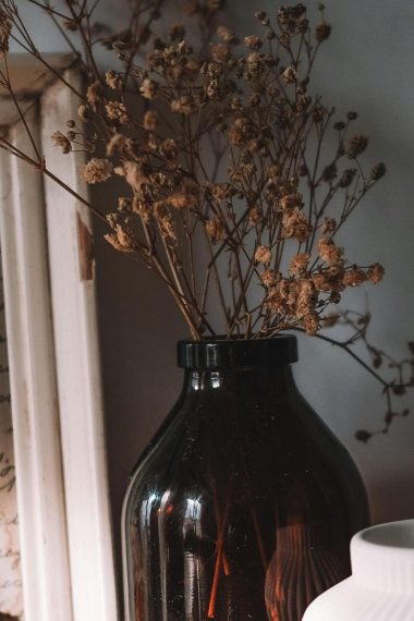 A close up of a dark red vase with dried flowers in it sitting on a table. against a concrete wall. A white vase in the foreground and picture frame in background.